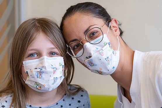 Das Bild ist eine Porträtaufnahme einer Ärztin und eines Mädchens, die beide bunte Masken tragen.