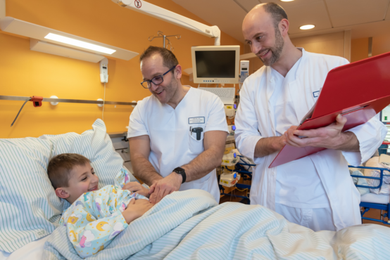 Das Bild zeigt den Leitenden Kinderchirurgen und einen Oberarzt der Kinderchirurgie auf Visite bei einem Jungen, der im Krankenhausbett liegt.